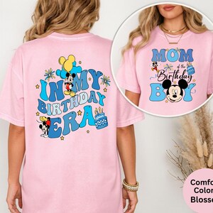 Op de afbeelding: Lichtroze t-shirt met de tekst "IN MY BIRTHDAY ERA" in blauw, met Mickey Mouse-afbeeldingen. De voorkant van het shirt heeft de tekst "MOM of the Birthday Boy" met Mickey Mouse-afbeeldingen. Onderdeel van de Comfort Colors Blossom collectie.