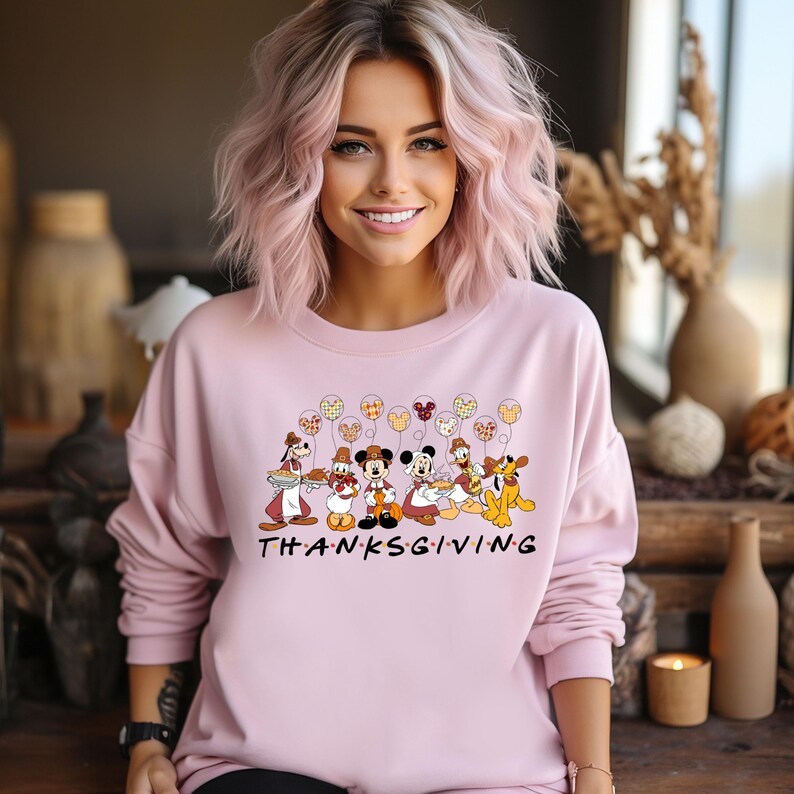 K&ouml;nnte beinhalten: Rosafarbener Pullover mit Thanksgiving-Motiv mit Mickey Mouse und seinen Freunden. Die Figuren halten Ballons mit Patchwork-Muster. Der Text "THANKSGIVING" ist unter den Figuren gedruckt.