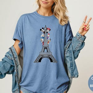 May include: Blue t-shirt featuring a black graphic of the Eiffel Tower with cartoon characters. The shirt has short sleeves and a crew neck. The text "Comfort Colors Blue Jean" is in a circle on the bottom right.