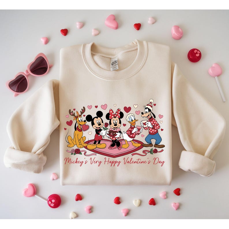 May include: Cream-colored sweatshirt featuring Mickey Mouse, Minnie Mouse, Donald Duck, Goofy, and Pluto, celebrating Valentine's Day. The graphic includes hearts, roses, and the text "Mickey's Very Happy Valentine's Day". Pink heart-shaped sunglasses and candies surround the sweatshirt.