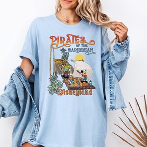 May include: Light blue t-shirt with a vintage-style graphic of the Pirates of the Caribbean ride at Disneyland. The graphic features Mickey Mouse and other Disney characters on a pirate ship.