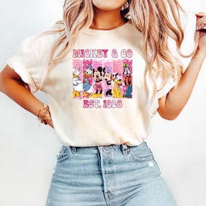 Puede incluir: Una camiseta blanca con un gr&aacute;fico rosa y blanco de Mickey Mouse y sus amigos. El gr&aacute;fico dice "Mickey & Co. Est. 1928".