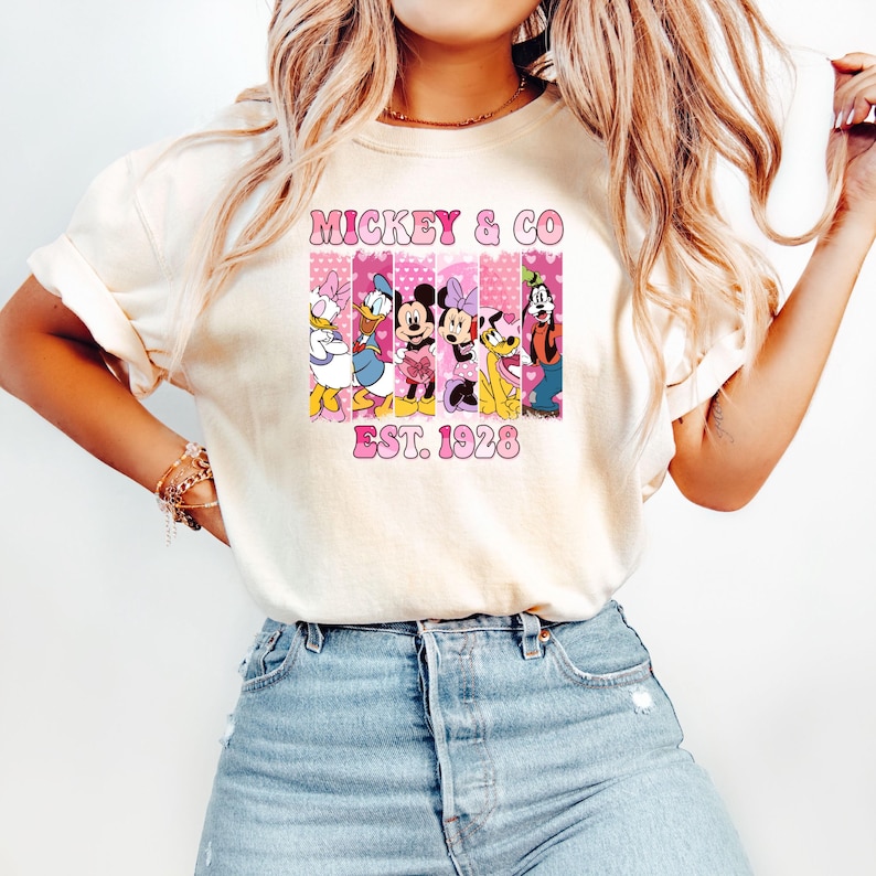 Puede incluir: Una camiseta blanca con un gr&aacute;fico rosa y blanco de Mickey Mouse y sus amigos. El gr&aacute;fico dice "Mickey & Co. Est. 1928".