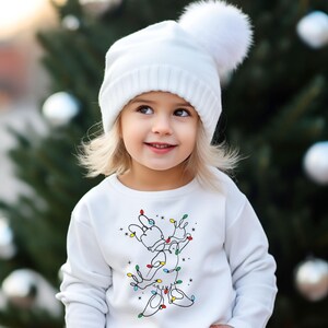 May include: A young child wearing a white knit hat with a large pom-pom and a white sweatshirt. The sweatshirt features a line drawing of a reindeer decorated with colorful Christmas lights. The background is a blurred Christmas tree.