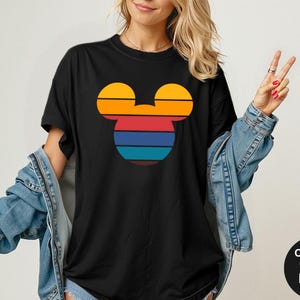 K&ouml;nnte beinhalten: Schwarzes T-Shirt mit einem Retro-Mickey-Mouse-Kopf-Design. Das Design zeigt horizontale Streifen in Orange, Rot, Blau und T&uuml;rkis. Das T-Shirt hat kurze &Auml;rmel und einen Rundhalsausschnitt. Der Text "Comfort Colors Black" befindet sich in einem schwarzen Kreis.