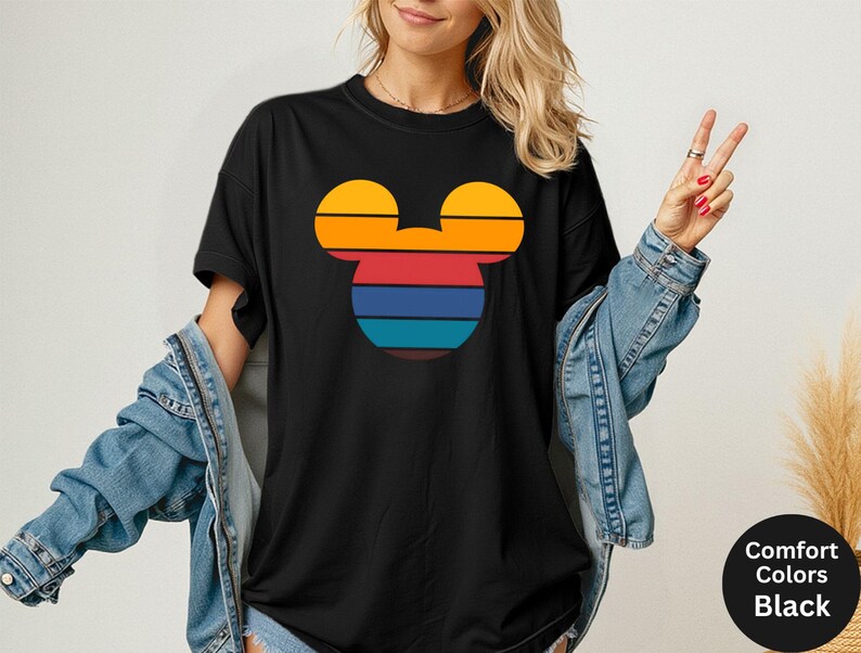 K&ouml;nnte beinhalten: Schwarzes T-Shirt mit einem Retro-Mickey-Mouse-Kopf-Design. Das Design zeigt horizontale Streifen in Orange, Rot, Blau und T&uuml;rkis. Das T-Shirt hat kurze &Auml;rmel und einen Rundhalsausschnitt. Der Text "Comfort Colors Black" befindet sich in einem schwarzen Kreis.