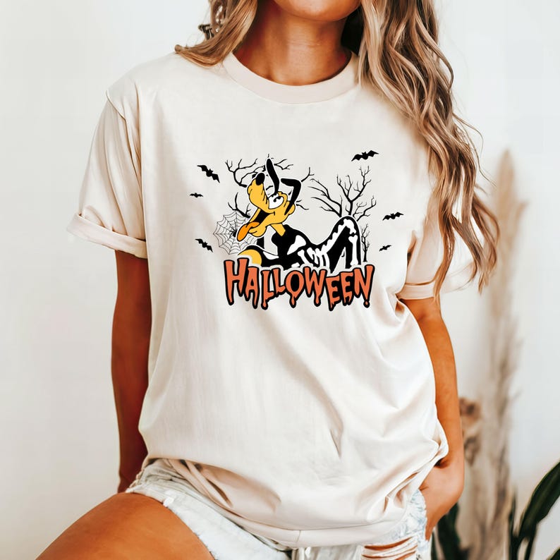 May include: Beige t-shirt featuring a Halloween-themed graphic. The design includes a cartoon dog in a Halloween costume, bats, a spider web, and the word "Halloween" in orange and black. The shirt has short sleeves and a crew neck.