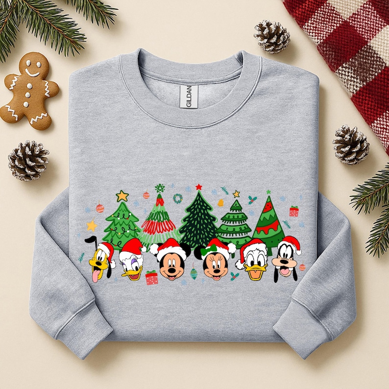 May include: A light gray sweatshirt featuring a festive design with cartoon characters wearing Santa hats and Christmas trees. The design includes Mickey Mouse, Minnie Mouse, Donald Duck, and Goofy. The sweatshirt is a Gildan brand.