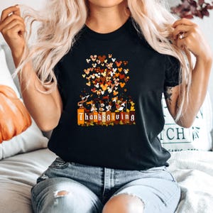 May include: Black t-shirt with a Thanksgiving-themed graphic. The design features a cluster of Mickey Mouse-shaped hearts in shades of brown, orange, and yellow. Below the hearts, there's a row of cartoon characters and the word "Thanksgiving".