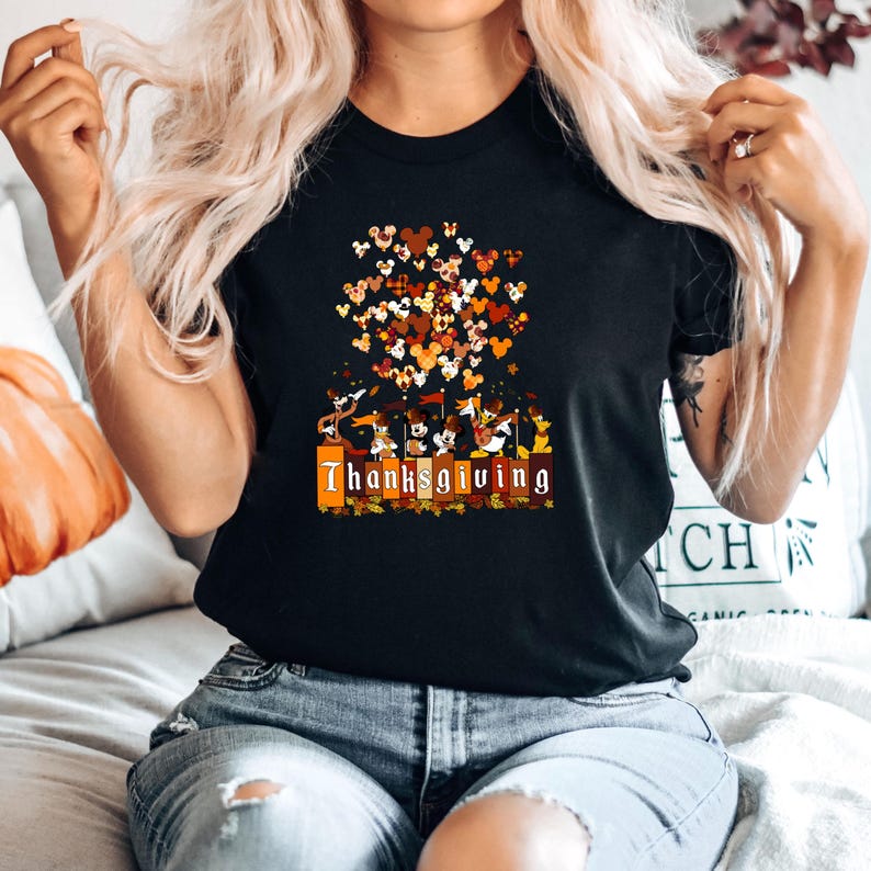 May include: Black t-shirt with a Thanksgiving-themed graphic. The design features a cluster of Mickey Mouse-shaped hearts in shades of brown, orange, and yellow. Below the hearts, there's a row of cartoon characters and the word "Thanksgiving".