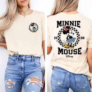 May include: A light beige t-shirt with a graphic of Minnie Mouse in a checkered circle with the text "Minnie Mouse" and "Disney" below. The graphic also includes the numbers "19" and "28".