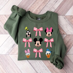 May include: A green sweatshirt with a graphic print of Mickey Mouse, Minnie Mouse, Donald Duck, and Goofy. Each character is wearing a pink bow.