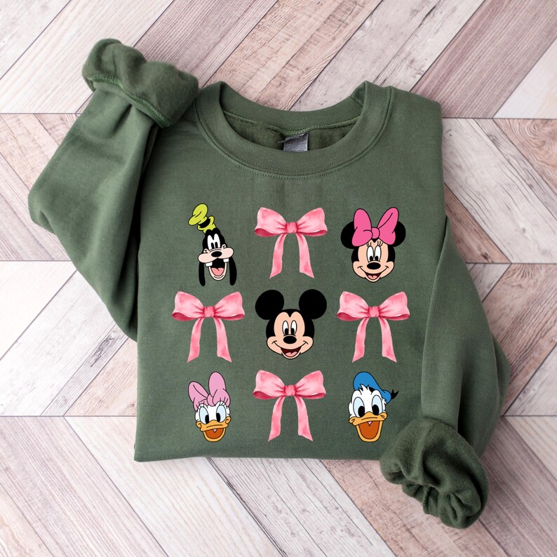 May include: A green sweatshirt with a graphic print of Mickey Mouse, Minnie Mouse, Donald Duck, and Goofy. Each character is wearing a pink bow.