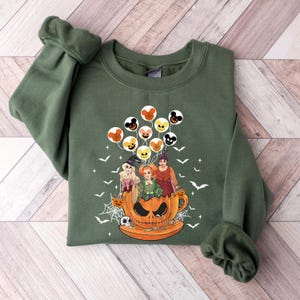 May include: Green sweatshirt with a graphic of three witches in a pumpkin teacup with Mickey Mouse shaped balloons. The witches are wearing Halloween costumes.