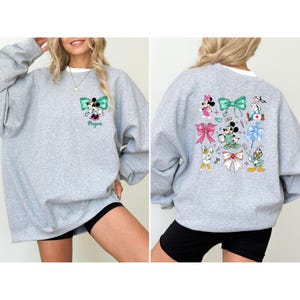 May include: A light gray sweatshirt featuring a cartoon design of Minnie Mouse and other Disney characters. The front has a teal bow and the name "Megan". The back has multiple bows and characters. The sweatshirt has a white collar.