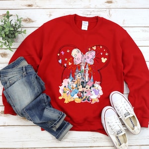 May include: Red sweatshirt with a graphic print of Mickey Mouse and friends in front of the Disney castle. The graphic also includes Minnie Mouse's iconic bow and hearts.