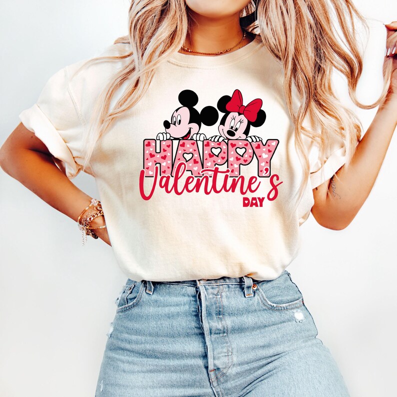 May include: White t-shirt with a pink and white heart pattern. The text "Happy Valentine's Day" is printed on the shirt with Mickey Mouse and Minnie Mouse peeking over the top of the letters.