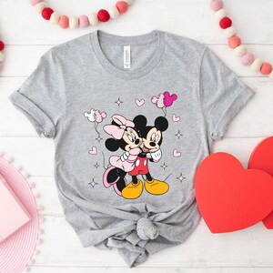 May include: A gray t-shirt with a graphic of Mickey Mouse and Minnie Mouse hugging. They are surrounded by pink hearts and stars. The text "CANVAS" is visible on the tag.