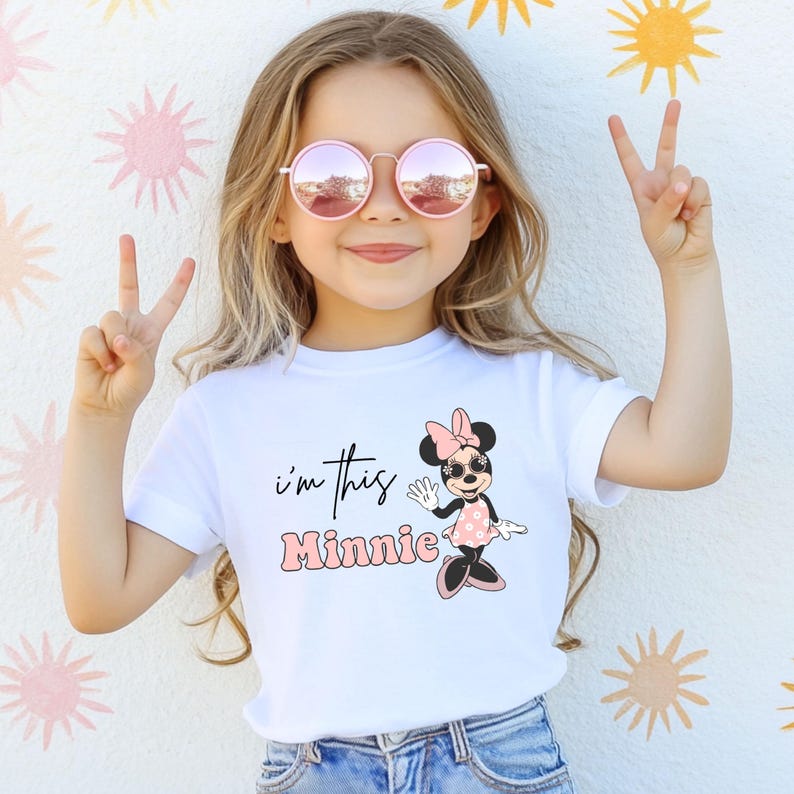 K&ouml;nnte beinhalten: Ein wei&szlig;es T-Shirt mit dem Text "I'm this Minnie" und einer Minnie Mouse-Cartoon-Grafik. Das Kind tr&auml;gt eine rosa Sonnenbrille und zeigt das Peace-Zeichen. Der Hintergrund hat ein Blumenmuster.