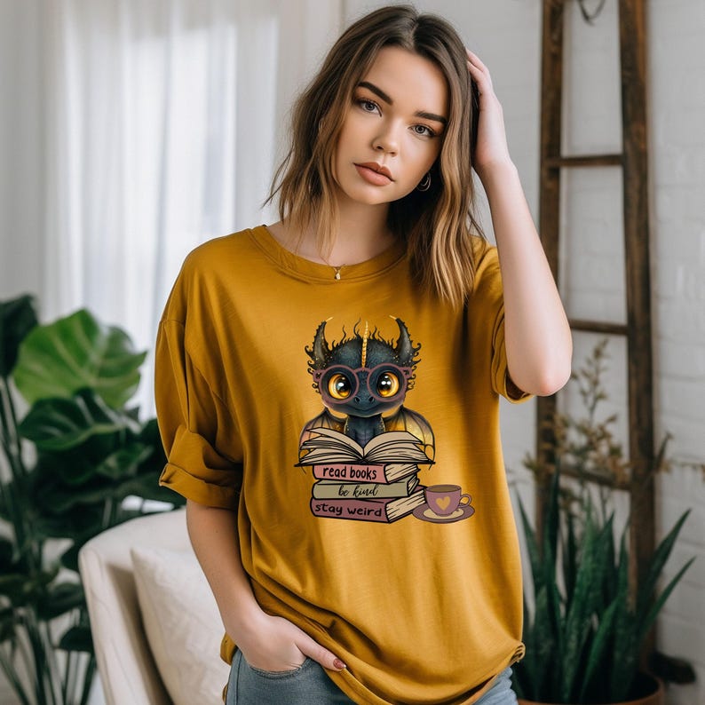 May include: A mustard yellow t-shirt with a cartoon dragon wearing glasses and holding a book. The text on the shirt reads "read books be kind stay weird".
