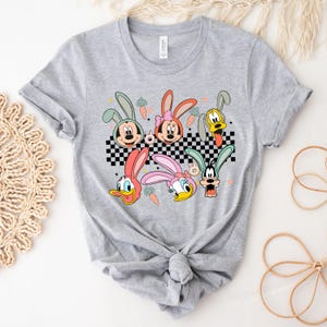 May include: A gray t-shirt with a checkered pattern and a graphic of cartoon characters with bunny ears. The characters include Mickey Mouse, Minnie Mouse, Donald Duck, Goofy, and Pluto.
