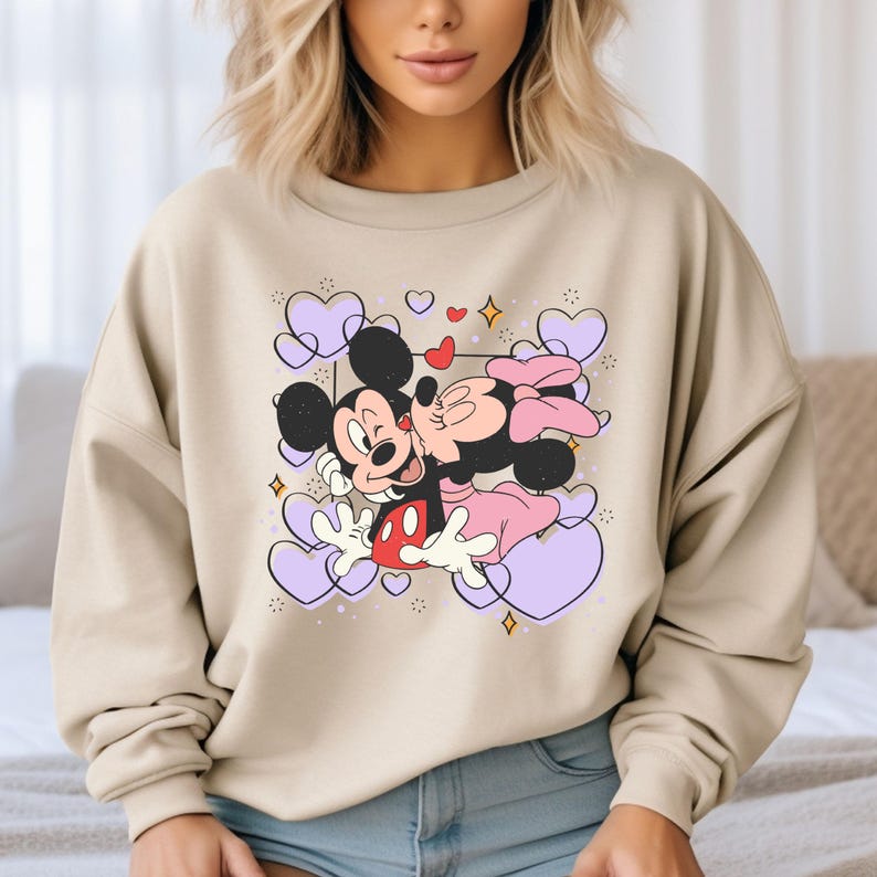 May include: A beige sweatshirt with a graphic print of Mickey Mouse and Minnie Mouse kissing. The graphic is surrounded by purple hearts and sparkles.