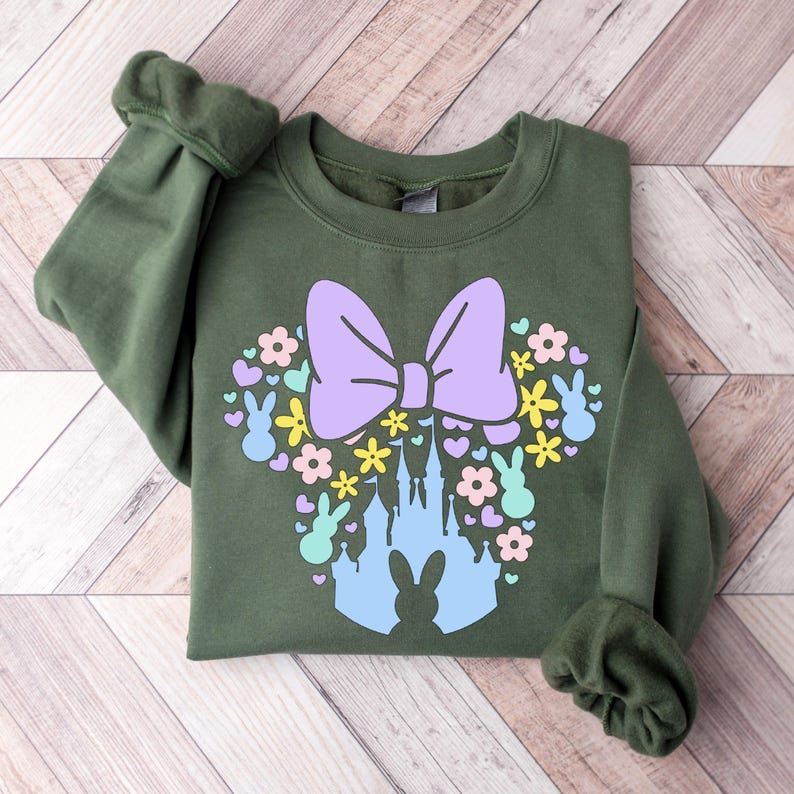 May include: An olive green sweatshirt featuring a colorful graphic. The graphic includes a large lavender bow, flowers, hearts, and bunny silhouettes arranged in a Mickey Mouse head shape. A light blue castle is in the center.