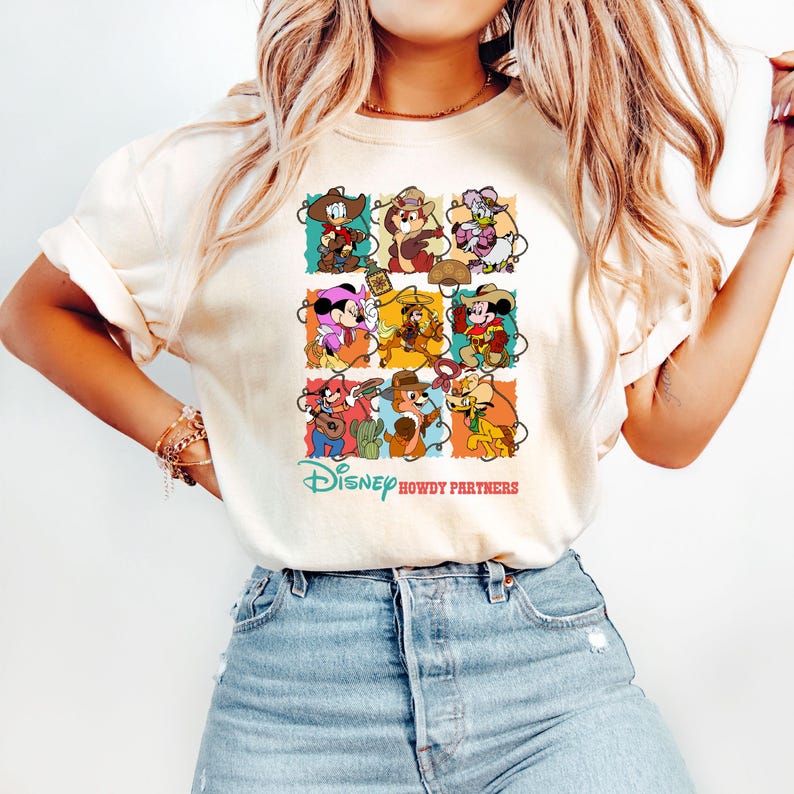 May include: Cream-colored t-shirt featuring a colorful graphic of Disney characters in cowboy attire. The design includes the text "Disney Howdy Partners" in a playful font, with various characters in a grid layout.