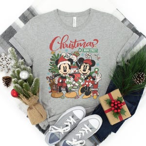 Op de afbeelding: Een grijs T-shirt met een kerstthema met Mickey en Minnie Mouse. Het ontwerp bevat een kerstboom, cadeaus en de tekst "Christmas on Main Street".