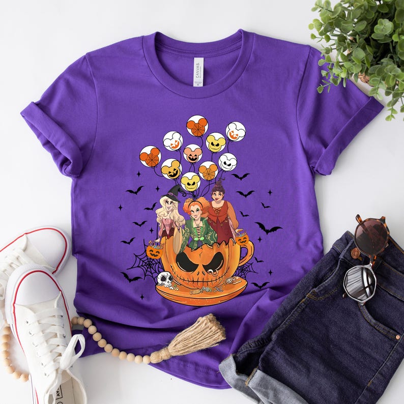 May include: Purple t-shirt with a graphic of three witches riding in a pumpkin-shaped teacup. The witches are wearing Halloween costumes and holding trick-or-treat bags.  Mickey Mouse-shaped balloons are floating above them.