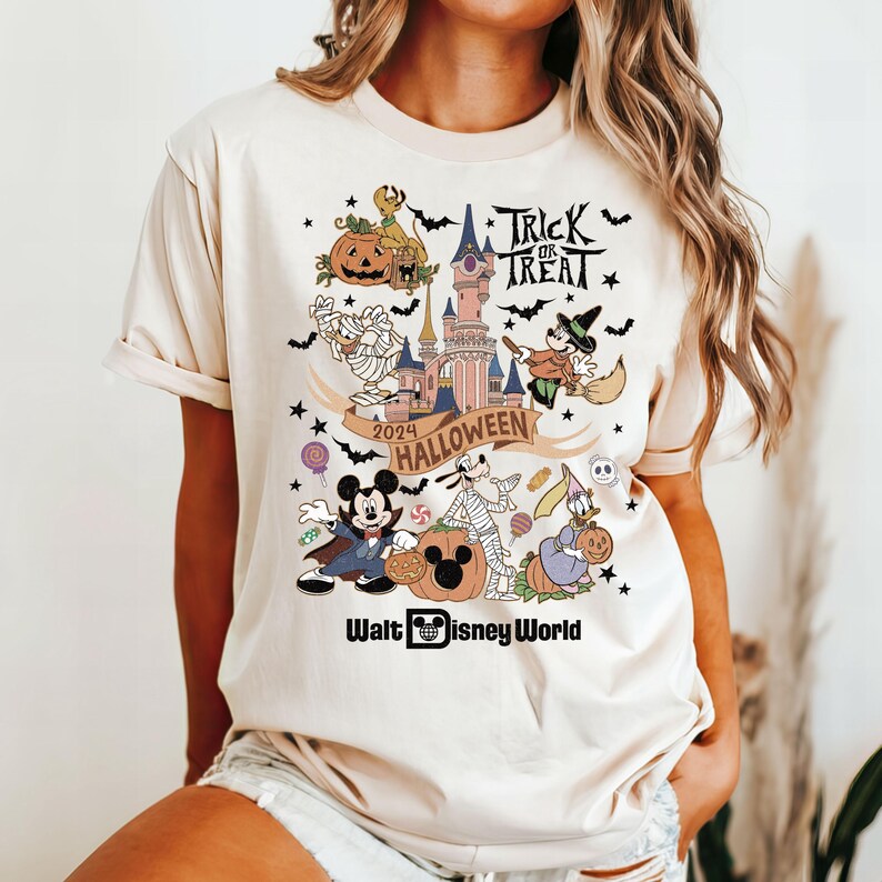 May include: A beige t-shirt with a colorful graphic print of Mickey Mouse, Minnie Mouse, Goofy, and other Disney characters celebrating Halloween at Walt Disney World. The graphic includes the text "Trick or Treat", "2024 Halloween", and "Walt Disney World".