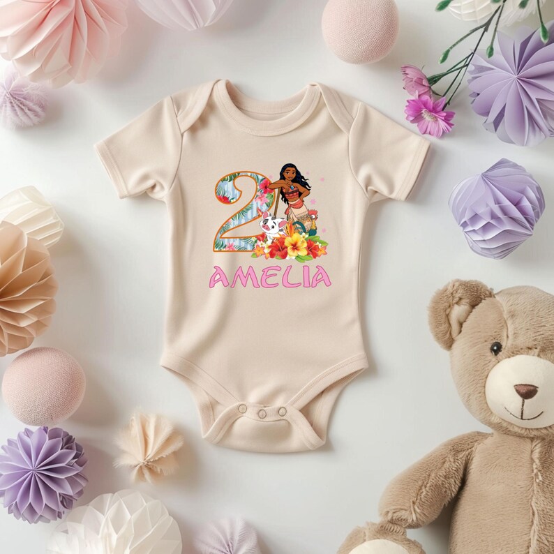 May include: A beige baby onesie with a colorful graphic featuring the number 2, a cartoon character, flowers, and the name "AMELIA" in pink. The onesie has short sleeves and snap closures.