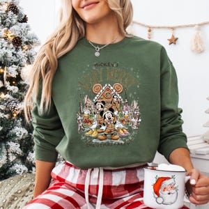Puede incluir: Sudadera verde con un dise&ntilde;o festivo de Mickey Mouse y sus amigos. El dise&ntilde;o incluye una casa de jengibre y el texto "Mickey's Very Merry Christmas Party". La sudadera se combina con un pantal&oacute;n de pijama a cuadros rojos y blancos.
