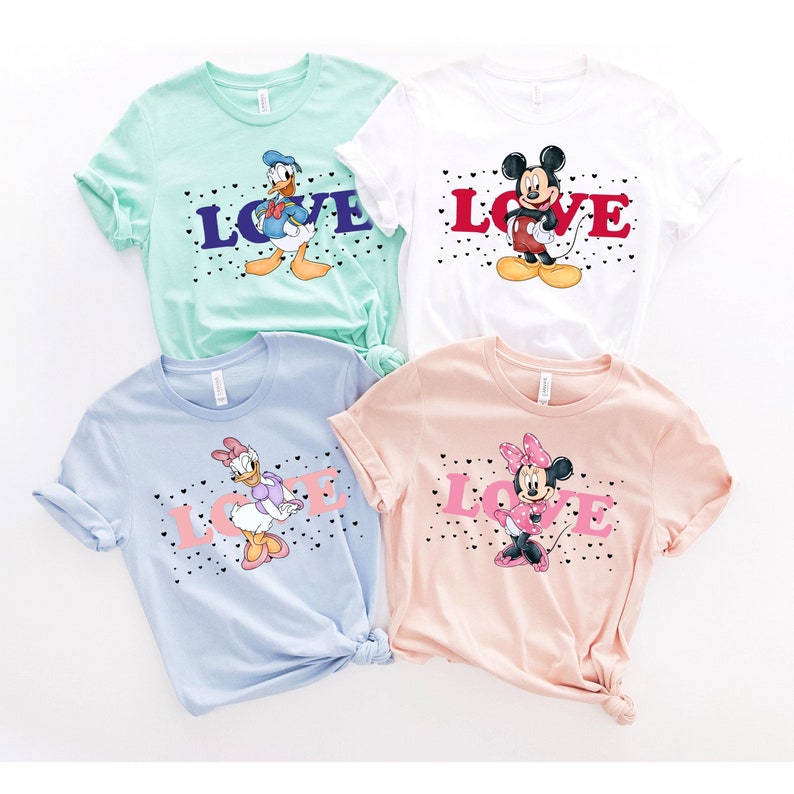 Op de afbeelding: Vier T-shirts met verschillende Disney-figuren. De shirts zijn allemaal wit, lichtblauw, lichtgroen en lichtroze. Elk shirt heeft een ander Disney-figuur erop, waaronder Mickey Mouse, Minnie Mouse, Donald Duck en Daisy Duck. De shirts hebben allemaal het woord "LOVE" in zwarte letters erop gedrukt.
