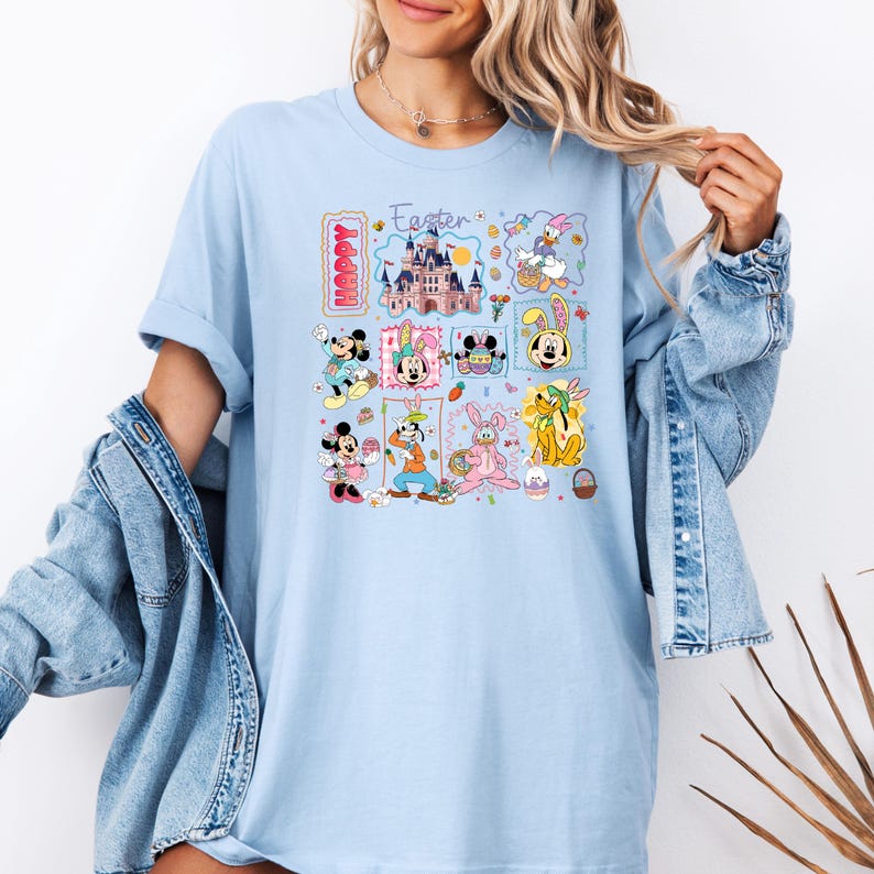 May include: Light blue t-shirt featuring a colorful graphic with Disney characters celebrating Easter. The design includes images of Mickey and Minnie Mouse, Donald Duck, and other characters, along with the words "Happy" and "Easter".