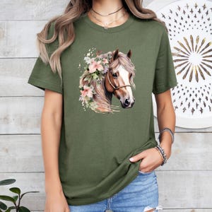 Peut inclure: T-shirt vert olive avec une illustration &agrave; l'aquarelle d'une t&ecirc;te de cheval orn&eacute;e de fleurs roses. Le cheval a une crini&egrave;re brune et un licol. Le t-shirt est de style d&eacute;contract&eacute;.