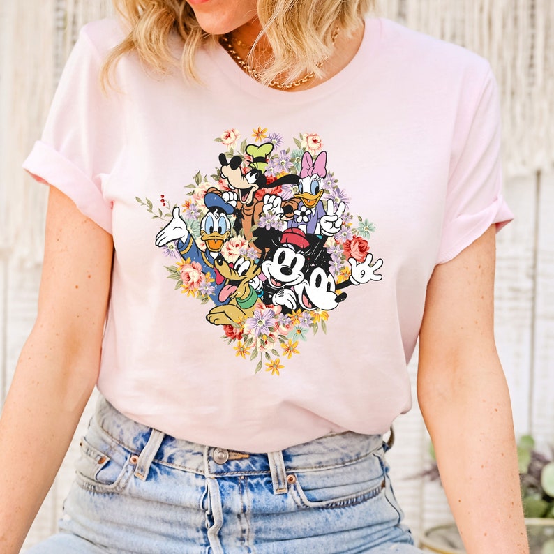 May include: Pink t-shirt with a colorful graphic print of Mickey Mouse, Minnie Mouse, Donald Duck, Goofy, and Pluto surrounded by flowers.