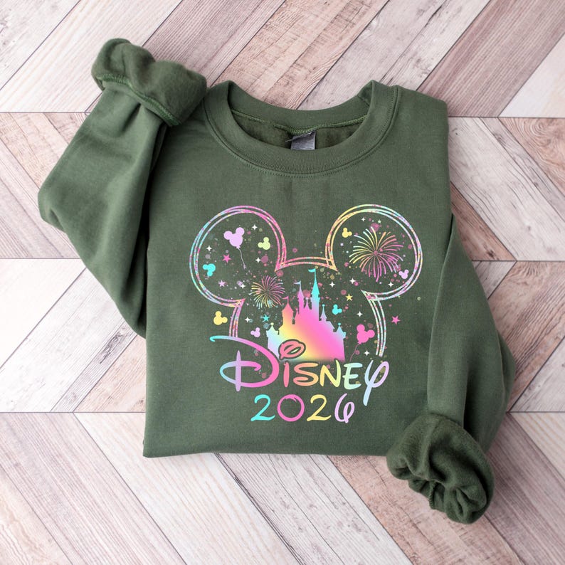 May include: An olive green sweatshirt featuring a colorful Disney design. The design includes a Mickey Mouse head outline with a castle, fireworks, and the text "Disney 2026" in a rainbow gradient.