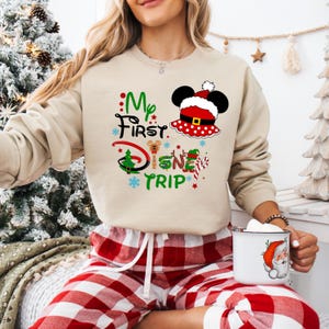 May include: A beige sweatshirt with a Christmas-themed design featuring Mickey Mouse wearing a Santa hat and the text "My First Disney Trip".