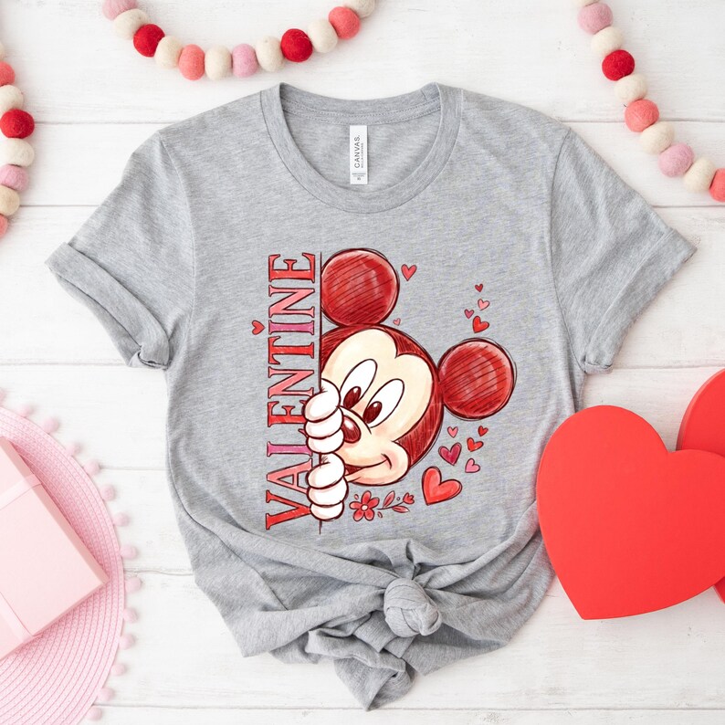 May include: A grey t-shirt with a cartoon Mickey Mouse design in red. The word "VALENTINE" is printed vertically in red. Small red hearts and flowers surround the character. The t-shirt is a casual style.
