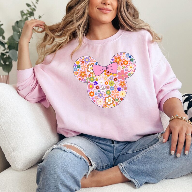 May include: A light pink sweatshirt featuring a floral Mickey Mouse design. The design includes a pink bow and the mouse's head and ears are filled with colorful flowers. The sweatshirt is a crew neck style.