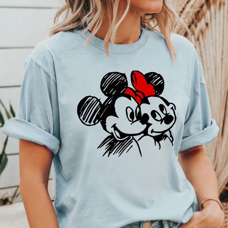 May include: Light blue t-shirt featuring a black and red graphic of Mickey and Minnie Mouse. The cartoon characters are smiling and embracing. Minnie Mouse is wearing a red bow. The shirt has rolled-up sleeves.