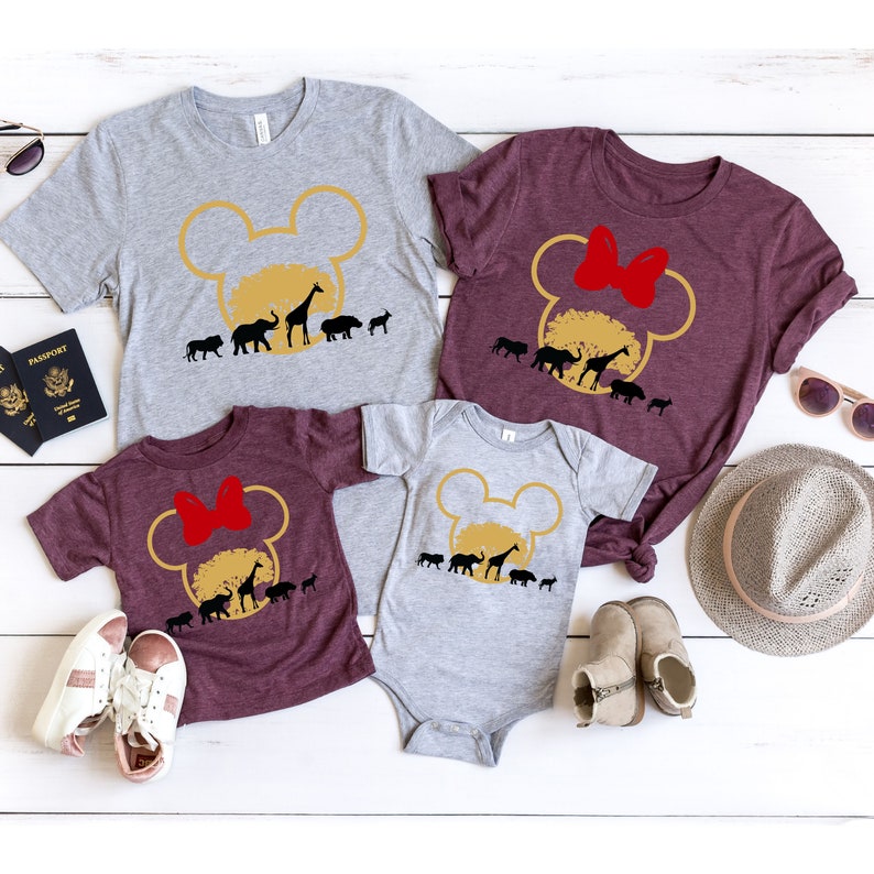 May include: A set of four t-shirts featuring a Mickey Mouse head silhouette with a safari scene inside. The shirts are in grey and maroon colors. The adult shirts have a short sleeve, while the children's shirts have a short sleeve and a long sleeve. The shirts are perfect for a family vacation to a safari park.