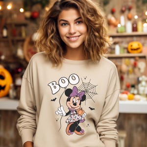 May include: A beige sweatshirt with a graphic print of Minnie Mouse dressed in a purple and black Halloween costume holding a pumpkin. The text "BOO" is printed in a bubble above her head.