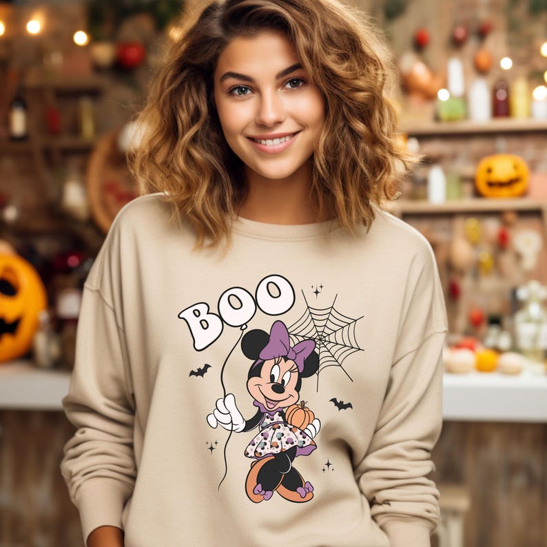 May include: A beige sweatshirt with a graphic print of Minnie Mouse dressed in a purple and black Halloween costume holding a pumpkin. The text "BOO" is printed in a bubble above her head.