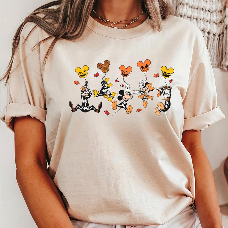 May include: Beige t-shirt with a Halloween graphic. The design shows cartoon characters in costumes, holding pumpkin-shaped balloons. The balloons are in different colours, and there are autumn leaves scattered around the characters.