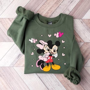 May include: Green sweatshirt with a graphic print of Mickey Mouse and Minnie Mouse hugging. The print includes pink hearts and balloons.