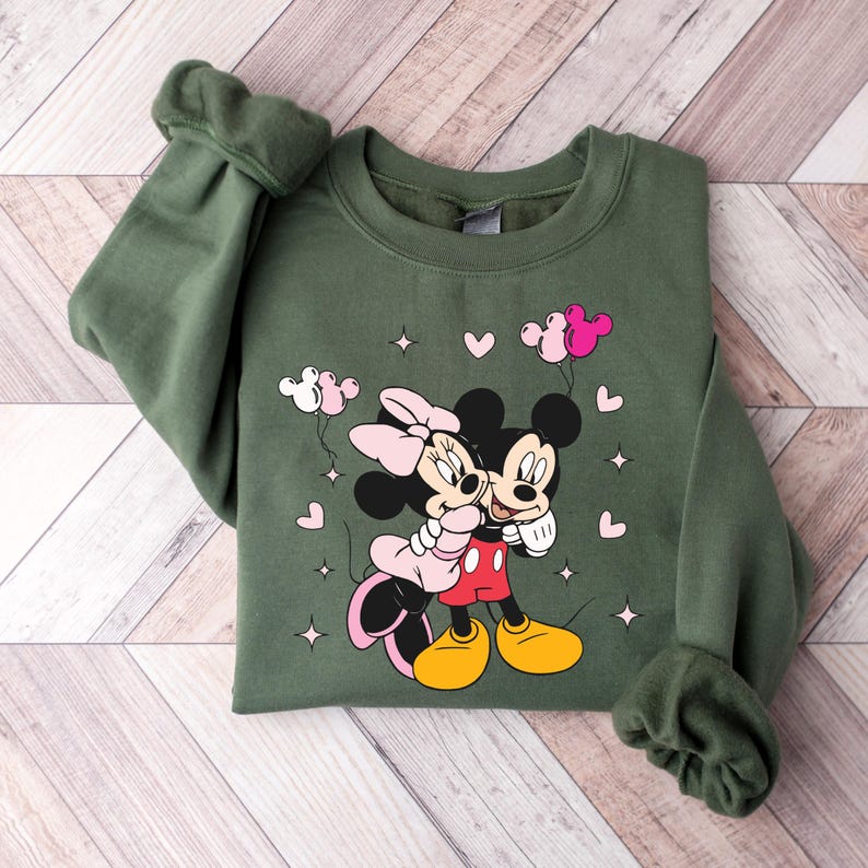 May include: Green sweatshirt with a graphic print of Mickey Mouse and Minnie Mouse hugging. The print includes pink hearts and balloons.