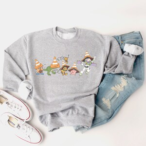 Op de afbeelding: Een grijs sweatshirt met een print van Toy Story-personages die verkeerskegels dragen. De personages zijn Woody, Buzz Lightyear, Mr. Potato Head, Rex en Hamm.