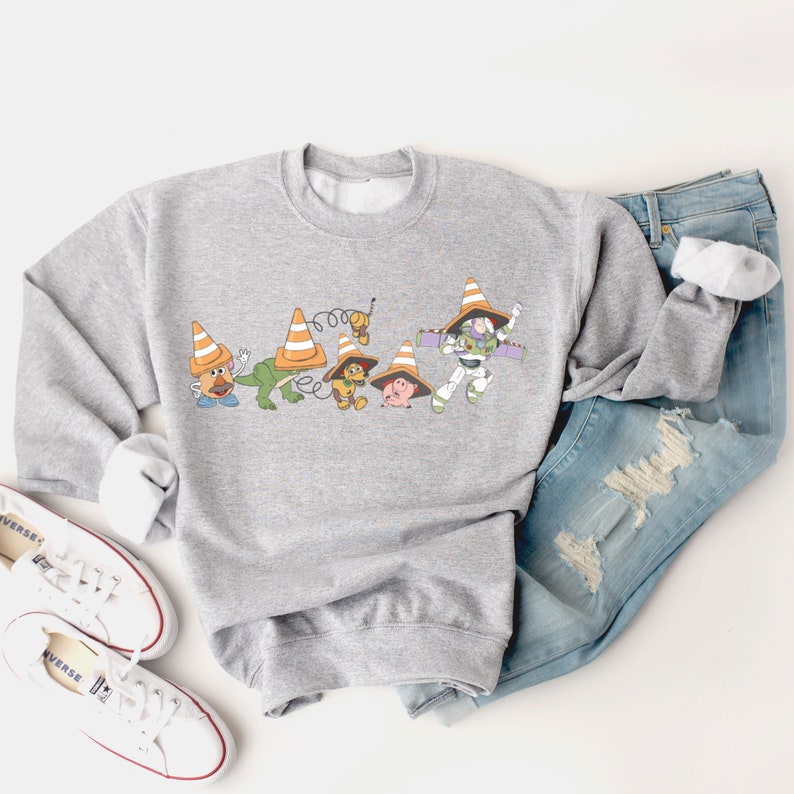 Op de afbeelding: Een grijs sweatshirt met een print van Toy Story-personages die verkeerskegels dragen. De personages zijn Woody, Buzz Lightyear, Mr. Potato Head, Rex en Hamm.